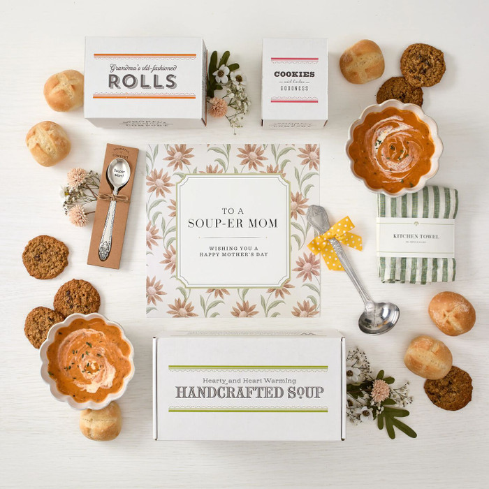 Overhead view of a Mother’s Day gift arrangement with two bowls of tomato soup, rolls, cookies, a box labeled “Handcrafted Soup,” a box of rolls, a box of cookies, a metal ladle with a yellow ribbon, a green striped towel, a packaged spoon, and a card that reads “To a Soup-er Mom."