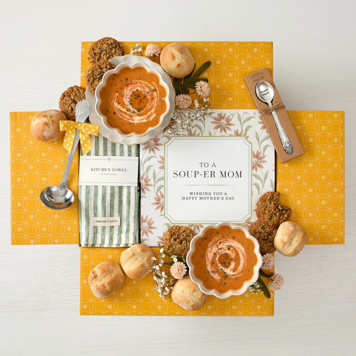 Overhead view of a yellow gift box arrangement with two bowls of soup, rolls, cookies, a metal ladle with a yellow ribbon, a green striped cloth, a packaged spoon, and a card that reads “To a Soup-er Mom. Wishing you a Happy Mother’s Day.”