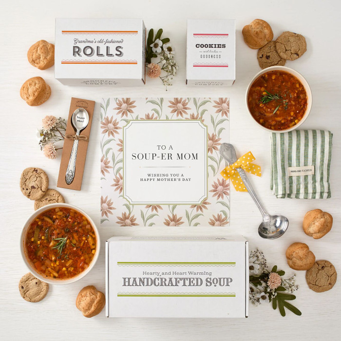Overhead view of a Mother’s Day gift arrangement with two bowls of tomato soup, rolls, cookies, a box labeled “Handcrafted Soup,” a box of rolls, a box of cookies, a metal ladle with a yellow ribbon, a green striped towel, a packaged spoon, and a card that reads “To a Soup-er Mom.