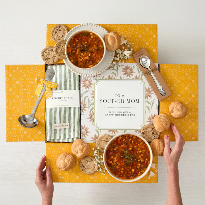 Overhead view of a yellow gift box arrangement with two bowls of soup, rolls, cookies, a metal ladle with a yellow ribbon, a green striped cloth, a packaged spoon, and a card that reads “To a Soup-er Mom. Wishing you a Happy Mother’s Day.”