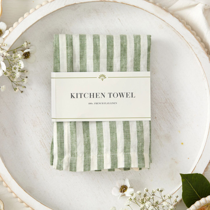 Close-up of a folded green striped kitchen towel wrapped with a label that reads “Kitchen Towel,” placed on a plate with small white flowers nearby.