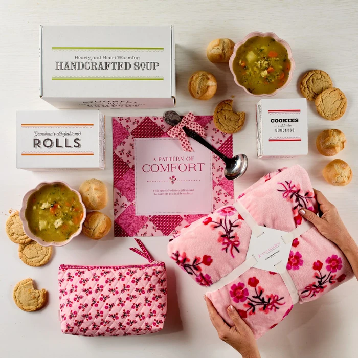 Cozy soup gift package arranged with bowls of soup, dinner rolls, cookies, a floral blanket, and pink floral accessories.