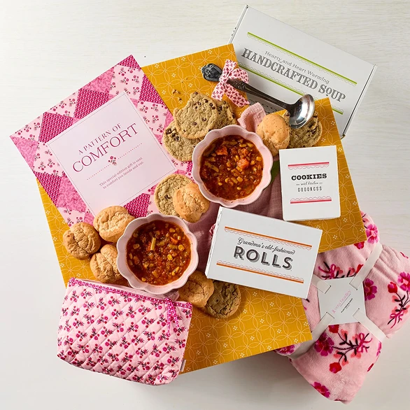 Overhead view of a comfort food gift package featuring two bowls of soup, dinner rolls, cookies, and a pink floral gift box.