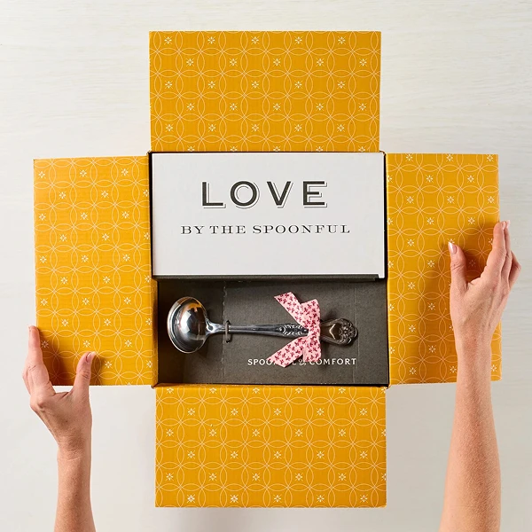 Hands opening a yellow patterned gift box featuring the word “Love by the Spoonful” on the lid and a small gift inside.