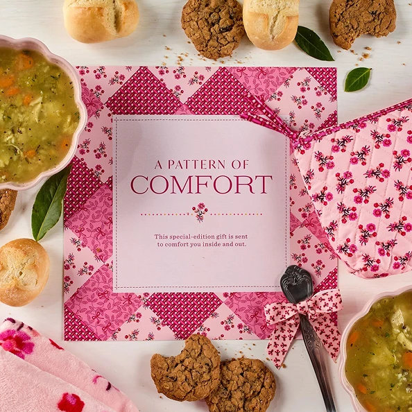 Overhead view of a pink floral comfort gift package surrounded by soup, dinner rolls, cookies, and decorative greenery.