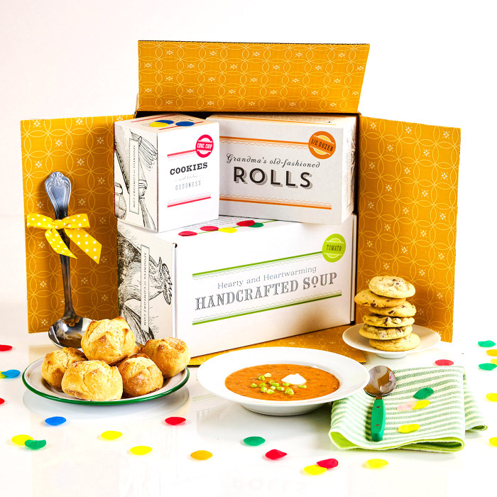 Open yellow gift box with packages labeled “Handcrafted Soup,” “Rolls,” and “Cookies,” surrounded by rolls, a bowl of soup, a stack of cookies, a green striped towel, a metal ladle with a yellow ribbon, and colorful confetti.