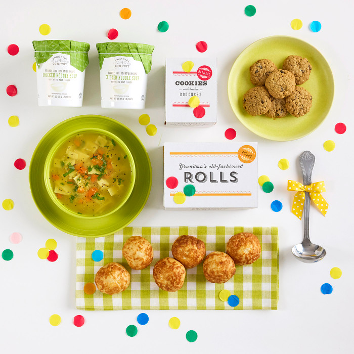 Overhead view of a bright arrangement with a bowl of soup, rolls, cookies, two containers of soup, a box of rolls, a metal ladle with a yellow ribbon, and colorful confetti scattered around.