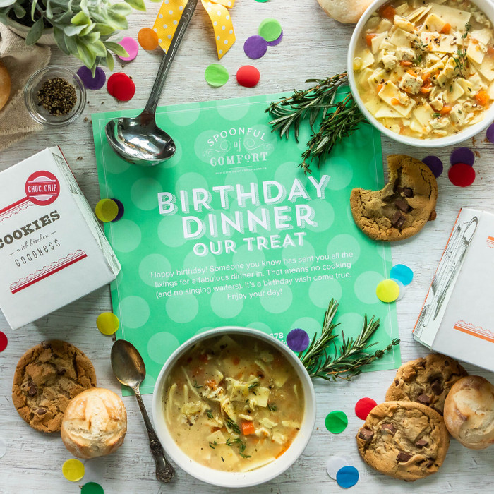 Overhead view of a birthday-themed food gift arrangement with bowls of soup, cookies, rolls, and packaged cookies surrounding a green card that reads “Birthday Dinner Our Treat,” with colorful confetti scattered on the table.
