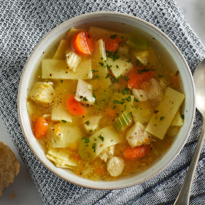 Close-up of a bowl of chicken noodle soup with carrots, celery, pasta, and herbs, placed on a gray cloth with a spoon nearby.