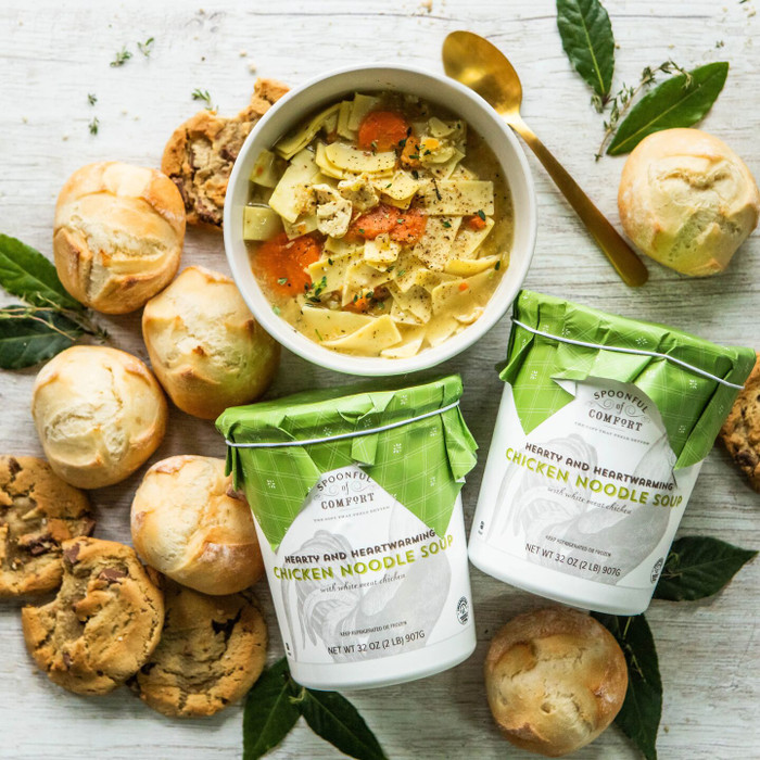 Overhead view of a bowl of chicken noodle soup surrounded by rolls, cookies, greenery, and two containers of soup with green lids.