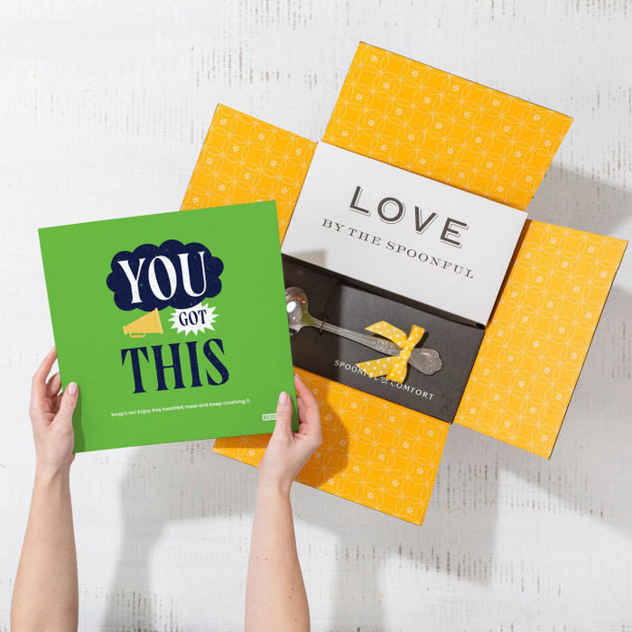 Overhead view of hands holding a green card that reads “You’ve Got This” next to an open yellow gift box with a card that reads “Love by the Spoonful” and a metal ladle with a yellow ribbon inside.