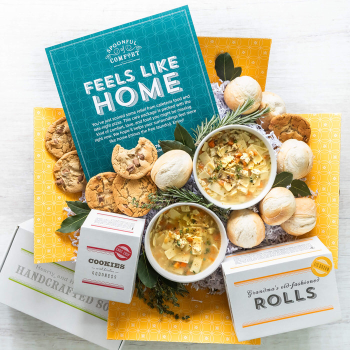 Unboxed College Care Package with an insert card with text that reads Feels Like Home. Displayed with two bowls of chicken noodle soup, chocolate chip cookies and dinner rolls.