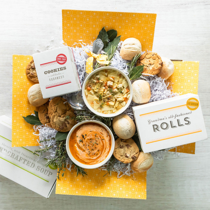 Overhead view of a yellow gift box arrangement with two bowls of soup, rolls, cookies, a container labeled “Handcrafted Soup,” and packaged rolls, arranged with greenery.