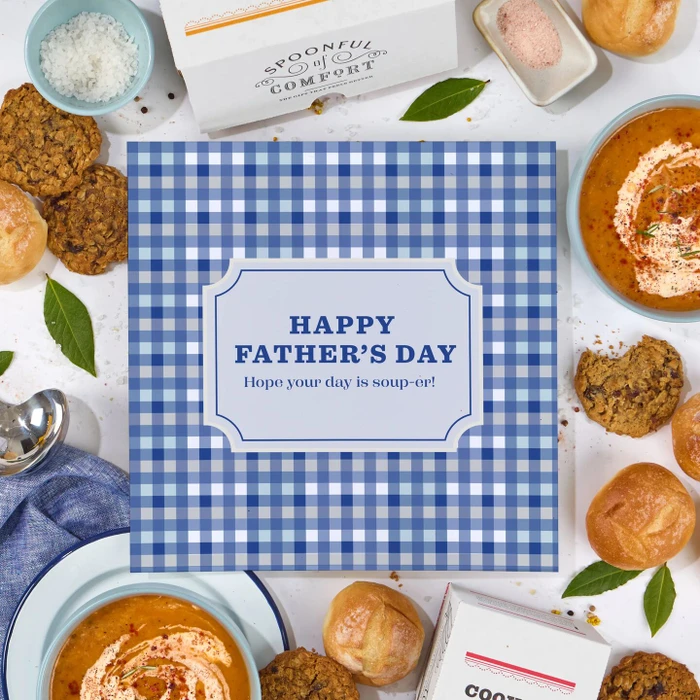 Close-up of a blue gingham card that reads “Happy Father’s Day. Hope your day is soup-er!” surrounded by bowls of soup, rolls, cookies, and greenery.