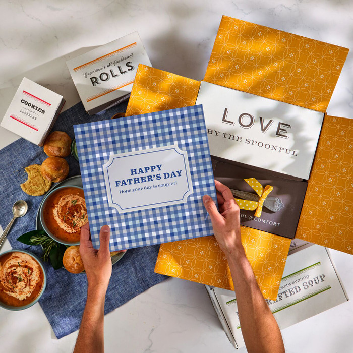 Overhead view of hands holding a blue gingham card that reads “Happy Father’s Day” above an open yellow gift box with a card that reads “Love by the Spoonful,” alongside bowls of soup, rolls, cookies, and a metal ladle with a yellow ribbon.