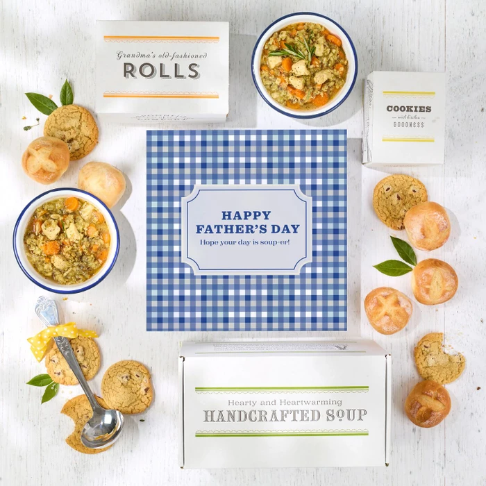 Overhead view of a Father’s Day gift arrangement with two bowls of soup, rolls, cookies, a box labeled “Handcrafted Soup,” a box of rolls, a box of cookies, a metal ladle with a yellow ribbon, greenery, and a blue gingham card that reads “Happy Father’s Day.”