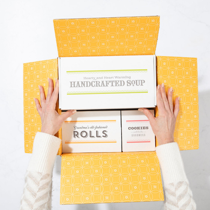 Top-down image of hands opening a gift box containing packaged handcrafted soup and bread rolls.