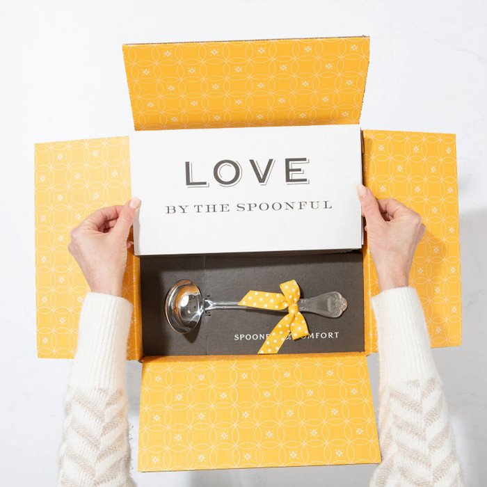 Overhead image of hands opening a gift box with branded packaging and a gold ribbon on a light background.
