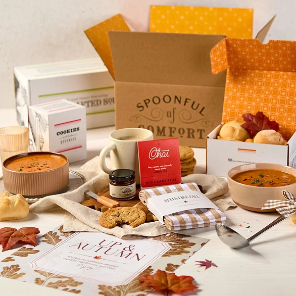 Overhead view of hands holding a fall-themed gift arrangement with two bowls of soup, rolls, cookies, a container labeled “Handcrafted Soup,” a metal ladle with a yellow ribbon, autumn leaves, and a card that reads “Up and Autumn.”