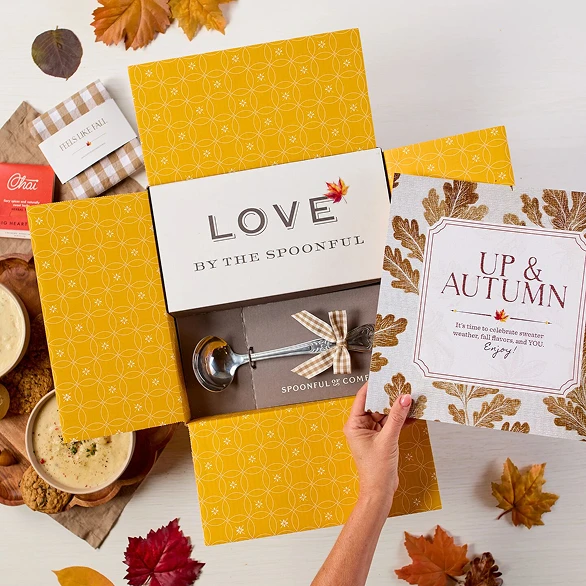 Overhead view of hands holding a fall-themed gift arrangement with two bowls of soup, rolls, cookies, a container labeled “Handcrafted Soup,” a metal ladle with a yellow ribbon, autumn leaves, and a card that reads “Up and Autumn.”
