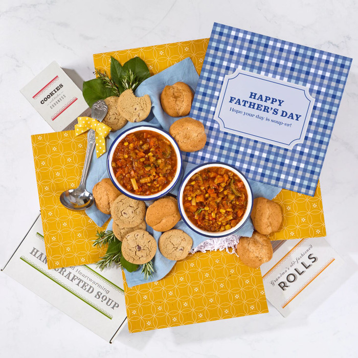 Overhead view of a Father’s Day gift arrangement with two bowls of soup, rolls, cookies, a container labeled “Handcrafted Soup,” a metal ladle with a yellow ribbon, and a blue checkered card that reads “Happy Father’s Day.”
