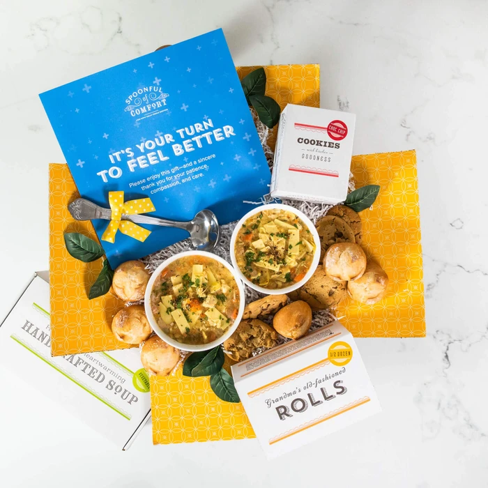 Overhead view of a yellow gift box arrangement with two bowls of soup, rolls, cookies, a container labeled “Handcrafted Soup,” a metal ladle with a yellow ribbon, and a blue card that reads “It’s Your Turn to Feel Better.”