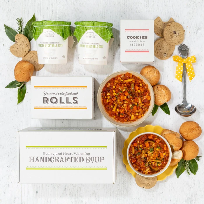 Overhead view of a soup gift arrangement with two bowls of soup, rolls, cookies, two containers of soup with green lids, a box labeled “Handcrafted Soup,” a box of rolls, a box of cookies, and a metal ladle with a yellow ribbon.