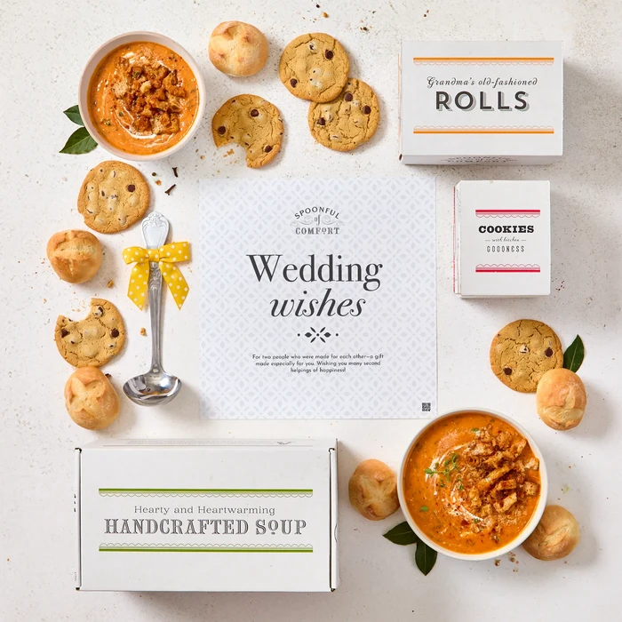 Overhead view of a wedding-themed gift arrangement with two bowls of soup, rolls, cookies, a box labeled “Handcrafted Soup,” a box of rolls, a box of cookies, a metal ladle with a yellow ribbon, and a card that reads “Wedding Wishes.”