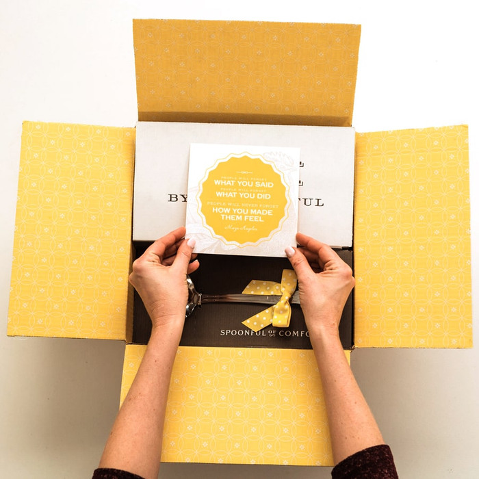 A Spoonful of Comfort shipping box opened to display a yellow printed interior. Hands reaching into a box holding an insert card that reads " people will forget what you said, people will forget what you did, but people will never forget how you made them feel.”
― Maya Angelou"