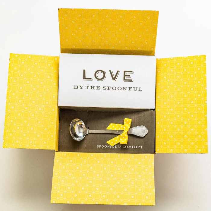 Open yellow gift box displaying a card that reads “Love by the Spoonful” and a metal ladle with a yellow ribbon inside.