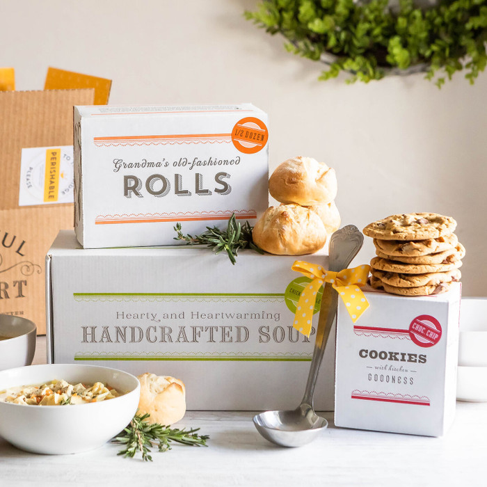 Gift set with packaged rolls, a container of handcrafted soup, a stack of cookies, and a bowl of soup. A metal ladle with a yellow ribbon tied to the handle is placed in front of the items.