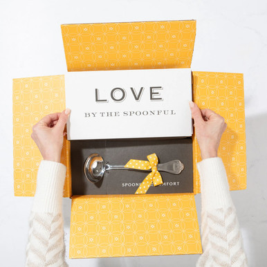 Open yellow gift box displaying a card that reads “Love by the Spoonful” and a metal ladle with a yellow ribbon inside.