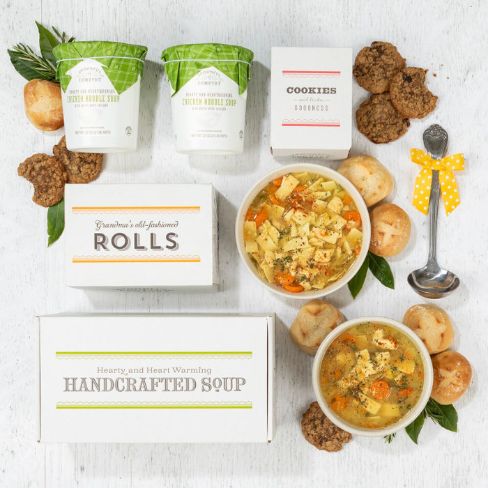 Overhead view of a soup gift arrangement with two bowls of chicken soup, rolls, cookies, two containers of soup with green lids, a box labeled “Handcrafted Soup,” a box of rolls, a box of cookies, and a metal ladle with a yellow ribbon.