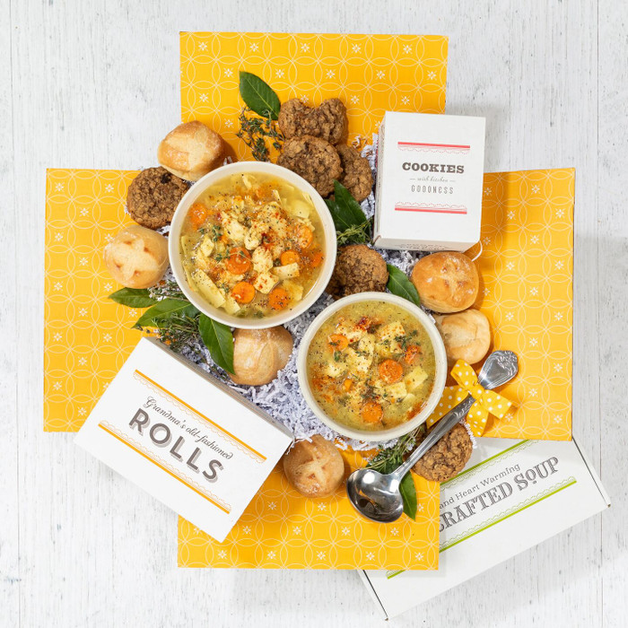 Overhead view of a yellow gift box arrangement with two bowls of chicken soup, rolls, cookies, a box of cookies, a box of rolls, a container labeled “Handcrafted Soup,” and a metal ladle arranged with greenery.