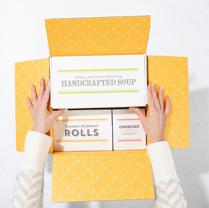 Overhead view of hands opening a yellow gift box containing packages labeled “Handcrafted Soup,” “Rolls,” and “Cookies.”