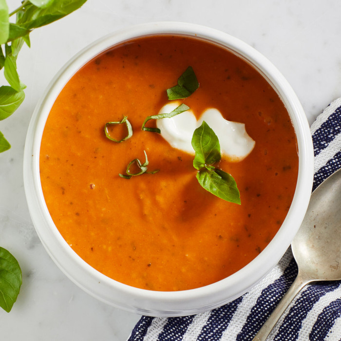 Close-up of a bowl of tomato soup garnished with cream and fresh basil, with a spoon and striped cloth nearby.