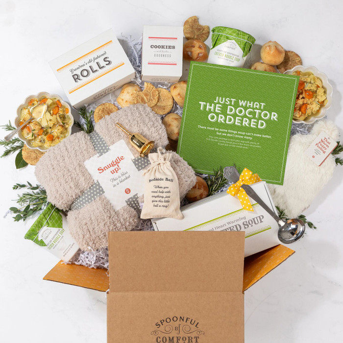Open Spoonful of Comfort gift box filled with a get-well-themed arrangement including two bowls of soup, rolls, cookies, containers of soup with green lids, a cozy throw blanket, a “Warm Fuzzies” warm/cool pack, a “Bedside Bell” for assistance, a metal ladle with a yellow ribbon, greenery, and a green card that reads “Just What the Doctor Ordered.”