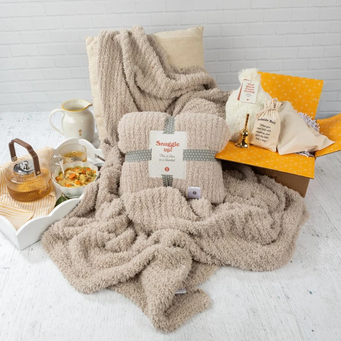 Cozy get-well-themed arrangement featuring a plush throw blanket with a “Snuggle Up” pack on top, alongside a tray with soup, tea, and honey, plus a “Bedside Bell” and self-care items displayed on a yellow box.