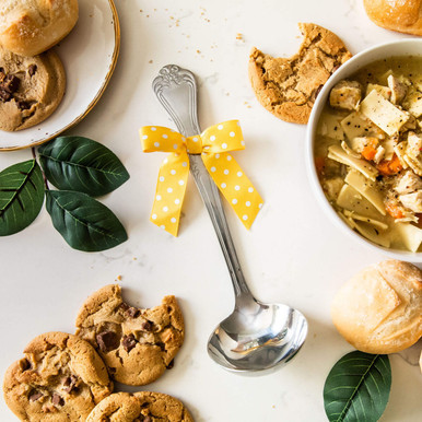 Overhead view of a metal ladle with a yellow ribbon surrounded by bowls of soup, rolls, cookies, and green leaves on a white surface.