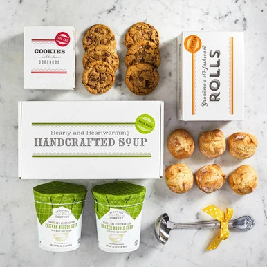 Overhead view of a soup gift set with a box labeled “Handcrafted Soup,” a box of rolls, cookies, two containers of soup with green lids, and a metal ladle with a yellow ribbon arranged on a surface.