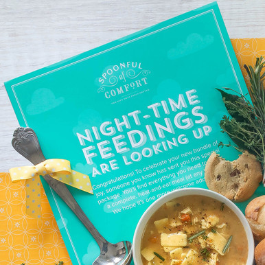 Close-up of a soup gift arrangement with a bowl of soup, rolls, a metal ladle with a yellow ribbon, and a teal card that reads “Night-Time Feedings Are Looking Up.”