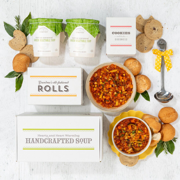 Overhead view of a soup gift arrangement with two bowls of chicken soup, rolls, cookies, two containers of soup with green lids, a box labeled “Handcrafted Soup,” a box of rolls, a box of cookies, and a metal ladle with a yellow ribbon.