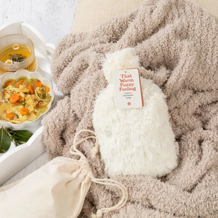 Close-up of a cozy plush blanket with a “Warm Fuzzies” warm/cool pack on top, next to a tray with a bowl of soup and a glass of tea.