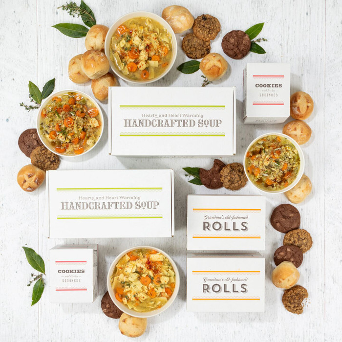 Overhead view of a symmetrical arrangement of soup gift items including four bowls of soup, rolls, cookies, multiple boxes labeled “Handcrafted Soup” and “Rolls,” and boxes of cookies arranged with greenery.