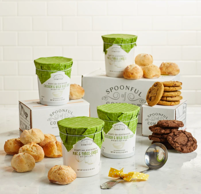 Soup gift set arranged on a counter with multiple containers of soup with green lids, rolls, cookies, Spoonful of Comfort boxes, and a metal ladle with a yellow ribbon.
