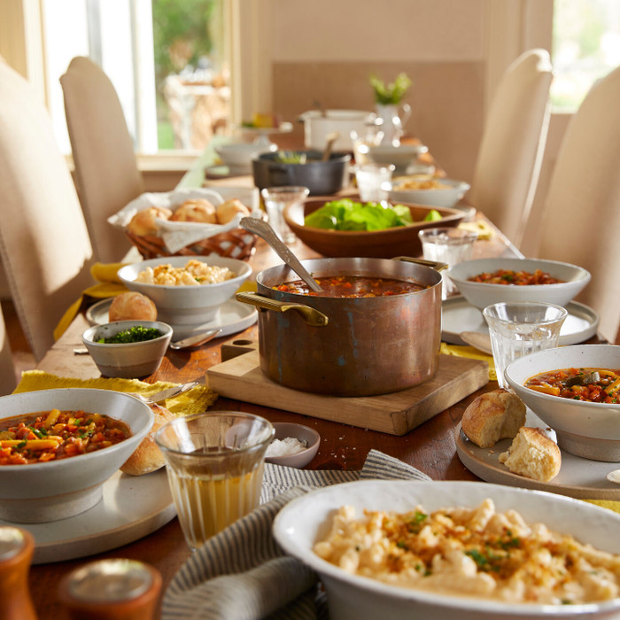 Dining table set with multiple bowls of soup, rolls, and drinks, centered around a pot of soup, with place settings arranged for a shared meal.