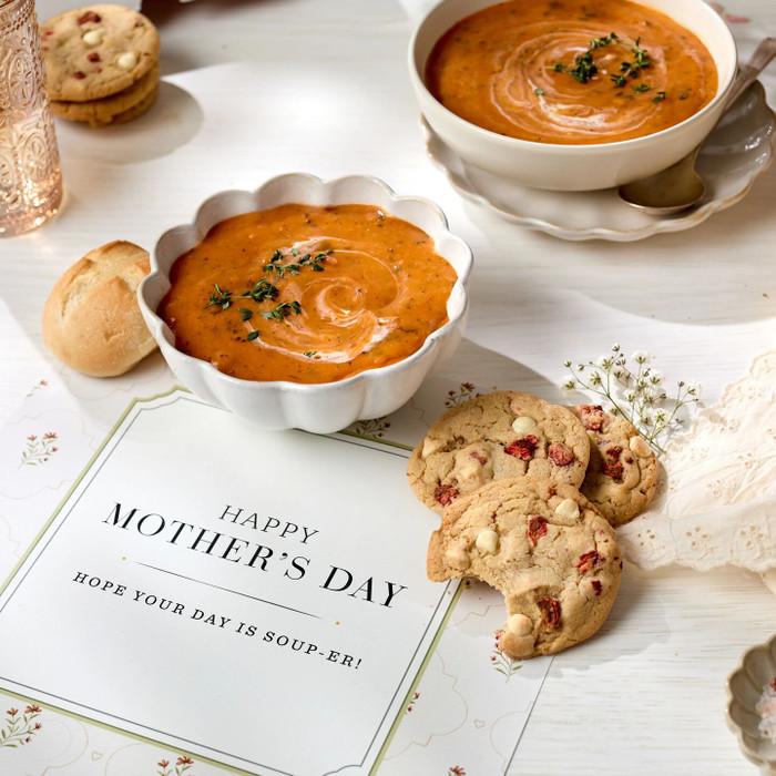 Close-up of a bowl of tomato soup with a roll and a partially eaten cookie beside a card that reads “Happy Mother’s Day. Hope your day is soup-er!”