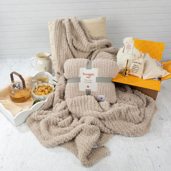Cozy get-well-themed arrangement featuring a plush throw blanket with a “Snuggle Up” pack on top, alongside a tray with soup, tea, and honey, plus a “Bedside Bell” and self-care items displayed on a yellow box.
