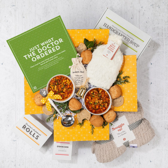 Overhead view of a yellow gift box filled with two bowls of soup, rolls, cookies, a container of handcrafted soup, a cozy throw blanket, a Warm Fuzzies warm/cool pack, and a green card that reads “Just What the Doctor Ordered.”