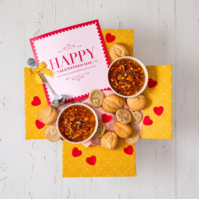 Overhead view of a Valentine’s-themed gift arrangement with two bowls of soup, rolls, cookies, a container labeled “Handcrafted Soup,” heart-shaped decorations, and cards that read “Happy Valentine’s Day”